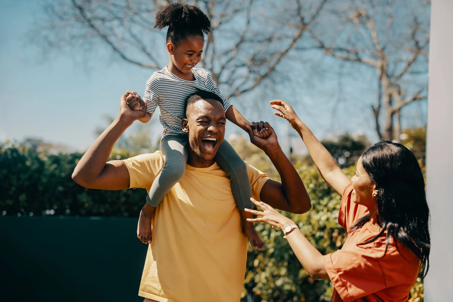 A child on an adult’s shoulders, sharing a playful moment with two adults outdoors.